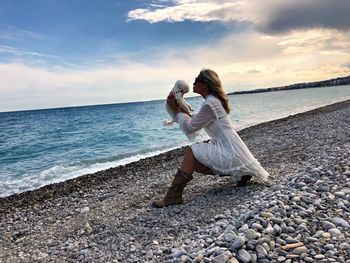 Woman wearing sunglasses with dog crouching at beach against sky