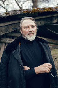 Elderly man with a beard stands in the village near a wooden shed in a sheepskin coat and glasses