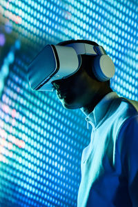 From below side view of concentrated young african american in white t shirt and modern vr goggles interacting with virtual object in dark studio with neon illumination