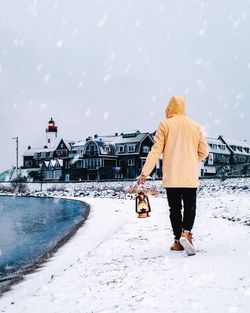 Rear view of man walking on shore against buildings during snowfall