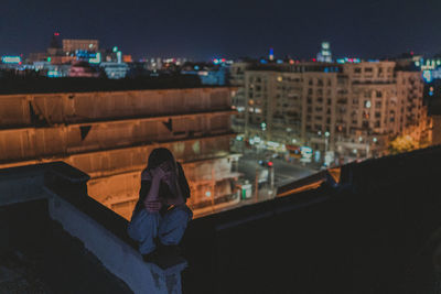 Woman sitting on retaining wall against buildings in city at night