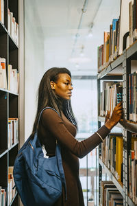 Side view of young woman with backpack searching book in library