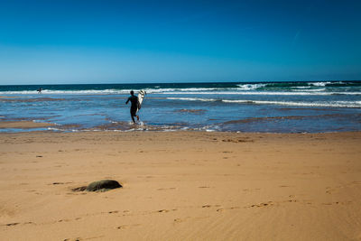 Rear view of man carrying surfboard while running towards sea on shore at beach