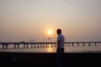 Side view of man standing by railing against sea during sunset