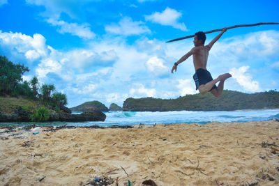 Full length of shirtless man holding stick while jumping at beach