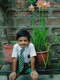 Portrait of cute boy wearing necktie sitting on railing by plant against wall