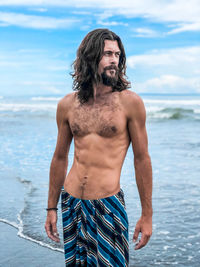 Shirtless man looking away standing at beach against sky