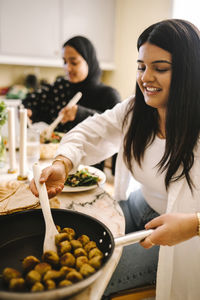 Smiling young woman serving food from cooking pan while sitting at dining table