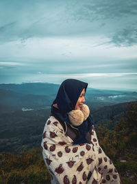 Woman wrapped in blanket looking away while standing against mountains