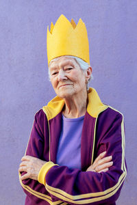 Confident elderly female athlete in sportswear and decorative crown looking at camera with folded arms on purple background