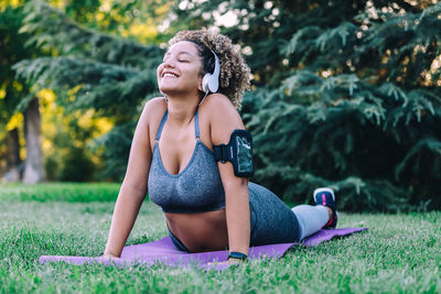 Optimistic young ethnic female in activewear listening to music with smartphone and headphones and doing back bend exercise during fitness training on green grass in park