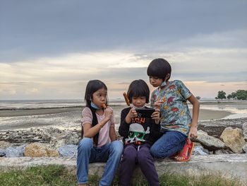 3 kids eating hotdog on stick, looking at smartphone isolated on beach background 