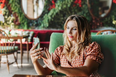 A young woman takes a selfie on her phone and sits at a table in a beautiful cafe