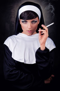 Portrait of nun smoking while standing against black background