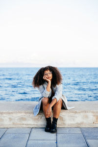 Young woman sitting on shore at sea against sky