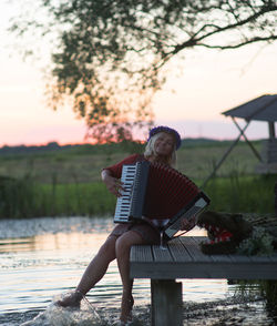 Rear view of man sitting by lake against sky during sunset