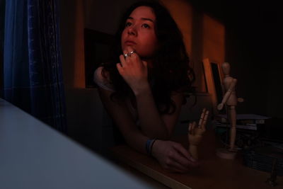 Young woman with hand on chin looking away while sitting by figurines on table at home