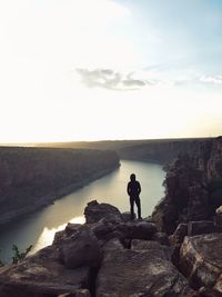 Man standing on rock while looking at river during sunset