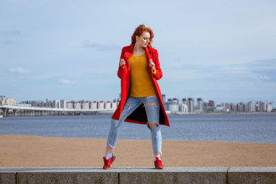 Red-haired woman poses against river in city with buildings in background.