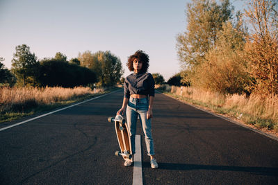 Portrait of young woman holding skateboard while standing on road during sunset