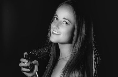 Portrait of smiling woman spraying perfume on neck against black background