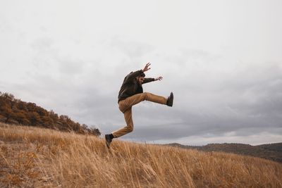 Full length of young man jumping on field against cloudy sky