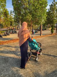 Rear view of woman walking with baby stroller on footpath at public park