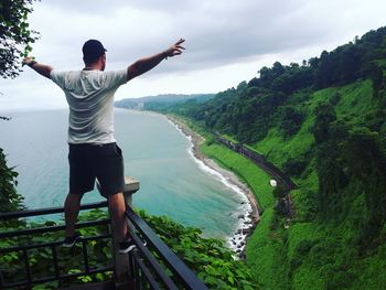 Rear view of man with arms outstretched standing at observation point by sea 