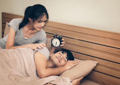 Woman holding alarm clock by men sleeping on bed at home