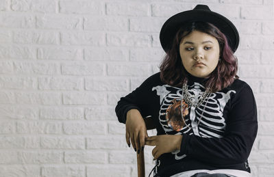 Young asian teen in costume witch sitting on a chair in front of a white brick wall,  halloween