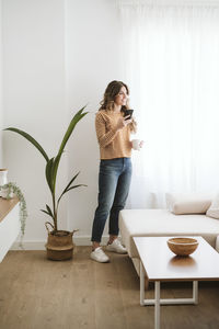 Smiling young woman holding coffee cup and mobile phone at home