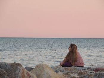 Side view of woman sitting at beach against clear sky during sunset