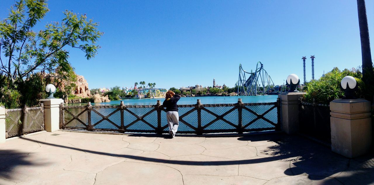 clear sky, lifestyles, leisure activity, water, blue, copy space, men, railing, full length, tree, person, sunlight, rear view, standing, built structure, sea, nature