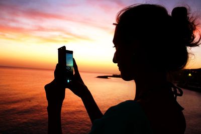 Side view of silhouette woman photographing sea against sky during sunset