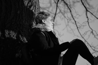 Side view of woman looking away while sitting against tree trunk