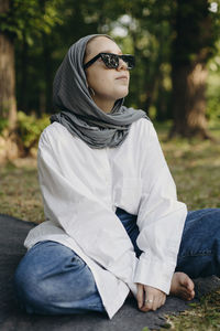 Young woman wearing sunglasses and hijab sitting cross-legged in park