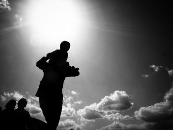 Low angle view of silhouette parent carrying child on shoulders against sky during sunny day