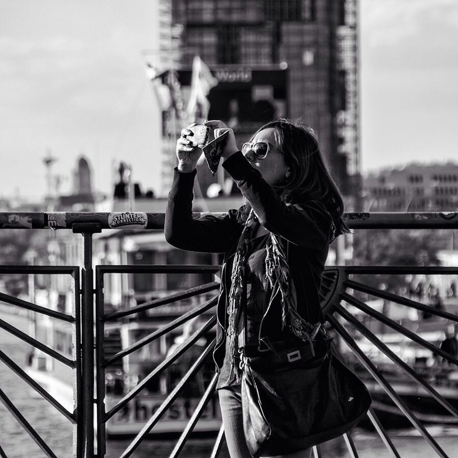 lifestyles, focus on foreground, leisure activity, young adult, casual clothing, standing, building exterior, three quarter length, built structure, person, holding, city, young women, architecture, railing, side view, sunglasses, rear view