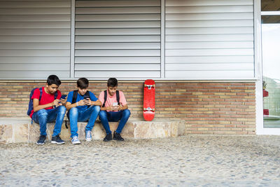Friends using smart phones while sitting against school building