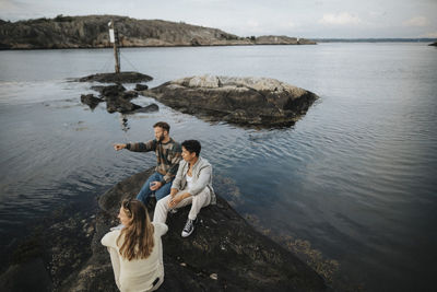 High angle view of man gesturing while sitting with friends on rock near lake