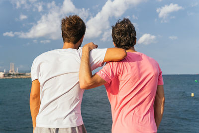 Back view of unrecognizable sportsmen leaning on each other and admiring sea and cloudy blue sky during break in fitness training on embankment