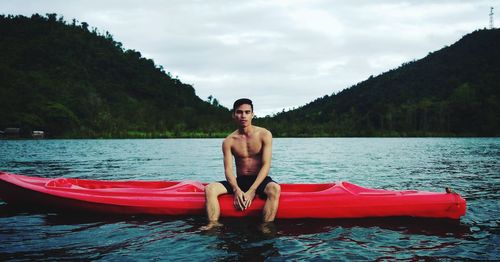 Portrait of shirtless man sitting in kayak on lake against mountains