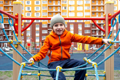 Funny caucasian blond boy in an orange jacket plays on the sports playground