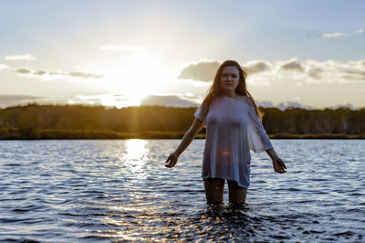 Portrait of young woman standing in lake against sky during sunset