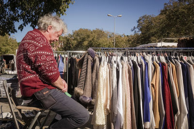 Senior man sitting by rack of clothes at flea market