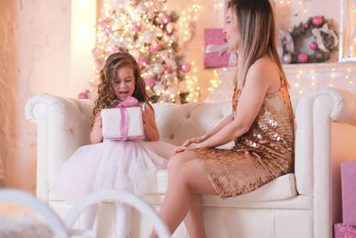 A fasion woman is presenting christmas gift for her daughter sitting on the sofa in light living 