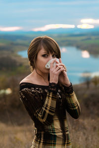 Portrait of young woman drinking while standing on mountain against cloudy sky