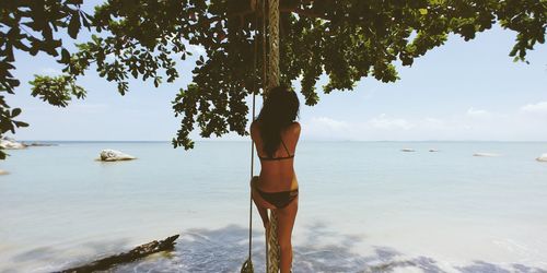 Rear view of woman wearing bikini while hanging on rope at beach