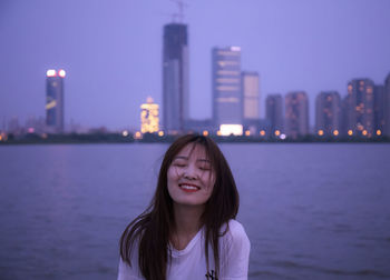 Smiling woman with eyes closed standing by river against illuminated buildings in city