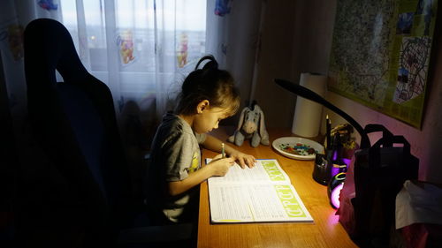 Side view of girl writing in book while sitting on table at home